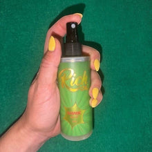 Load image into Gallery viewer, The Rich Oil Atomic Oil natural sun maximizer tanning oil in a green spray bottle held by a hand with yellow nails.